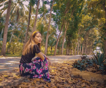 Portrait of young woman sitting on footpath against trees