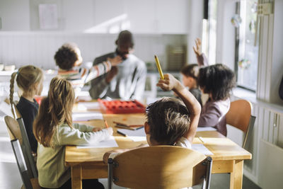 Boy raising hand while sitting in classroom during q and a session at kindergarten