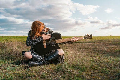 Woman sitting in a green field dressed in cowboy boots looking at a guitar under a sky with clouds