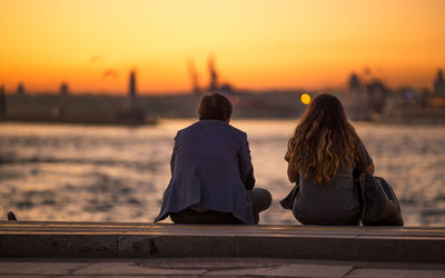 Rear view of couple sitting on pier against orange sky during sunset