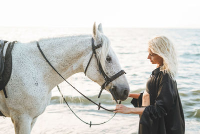 Young blonde woman in black clothes riding white horse on seascape background