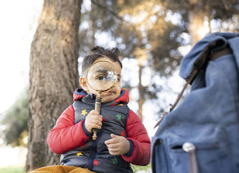 Boy in padded jacket looking through magnifying glass while sitting in park