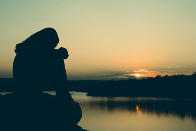 Silhouette woman sitting on rock at lakeshore sky during sunset