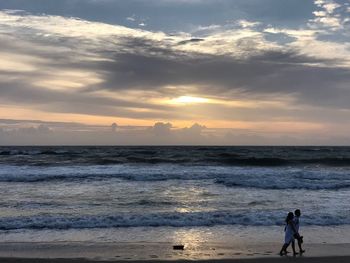 Silhouette people walking at beach against cloudy sky during sunset