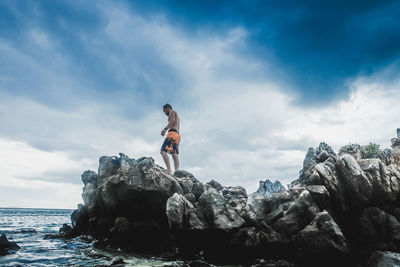 Low angle view of shirtless man standing on rock by sea against cloudy sky