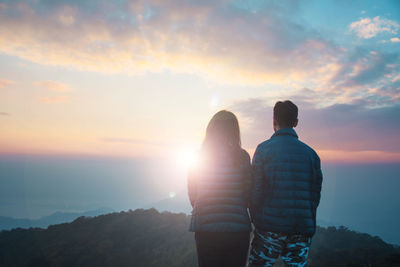 Rear view of couple standing on mountain against cloudy sky during sunset