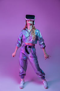 Full body of cheerful female with long hair in trendy outfit and vr glasses dancing in studio with purple neon illumination