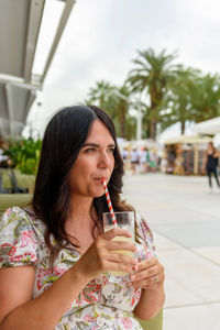 Pretty girl with long hair sitting in bar outdoor in city, drinking lemonade on straw