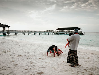 Father carrying daughter while looking at children playing on sand against sky