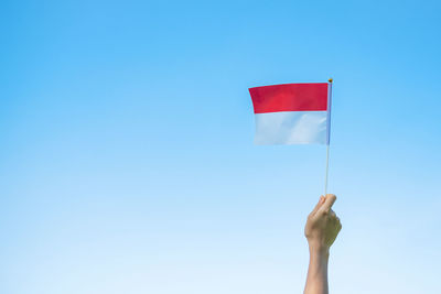 Low angle view of hand holding flag against clear blue sky