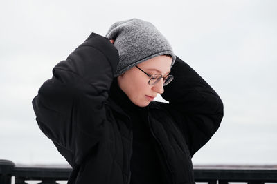 Young woman in hat, eyeglasses, black coat with hands up touching head looking down