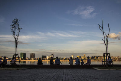 People on footpath by cityscape against sky at sunset