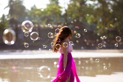 Side view of woman surrounded with bubbles while standing by pond