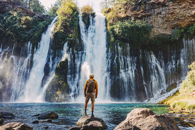 Man in a hat and yellow jacket looking on the beautiful waterfall, view from the back