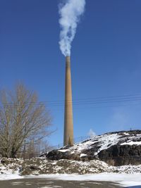 Low angle view of smoke stack emitting pollution against blue sky during winter