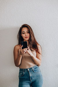 Young woman using phone while standing against wall