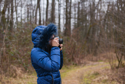 Side view of person photographing with camera while standing against bare trees