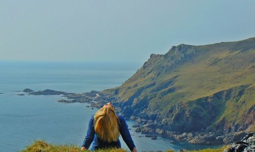 Rear view of woman tossing hair on cliff by sea against clear sky