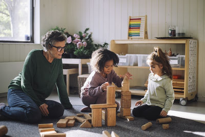 Senior female teacher sitting next to girls playing with toy blocks while sitting in classroom at kindergarten