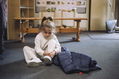 Girl tying shoelace while sitting with jacket on floor in classroom at kindergarten