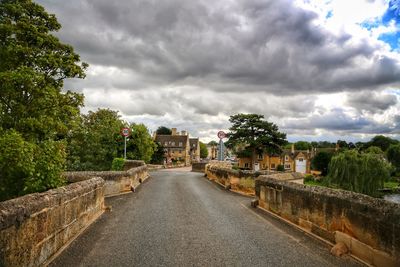 Road amidst trees and buildings against cloudy sky