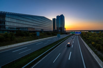 Road by buildings against sky during sunset in city