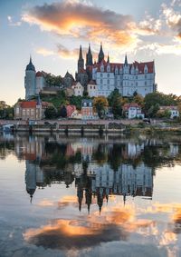 Buildings in town by river against sky during sunset