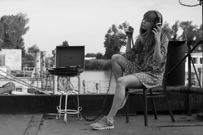 Woman listening music while sitting on chair against clear sky