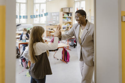 Happy teacher giving high-five to schoolgirl standing at doorway in school