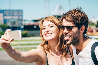 Happy friends clicking selfie on smart phone while standing outdoors