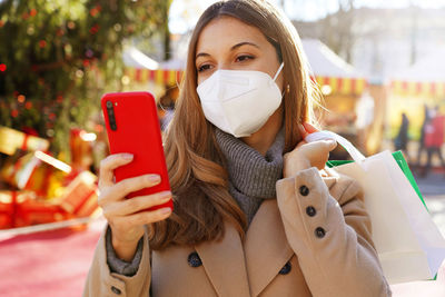 Young fashion woman with kn95 ffp2 face mask watching her telephone walking between christmas market