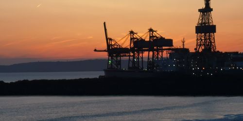 Silhouette cranes at commercial dock against sky during sunset