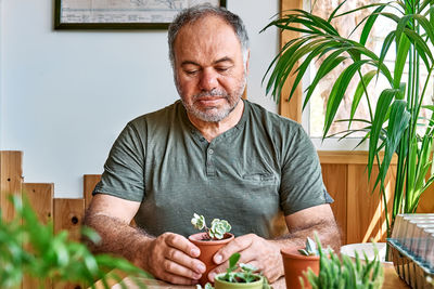 Mature bearded man taking care of plants and home flowers, planting  succulent in pot on the table.