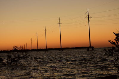 Silhouette electricity pylons by sea against orange sky
