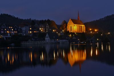 Illuminated house by lake against sky at dusk