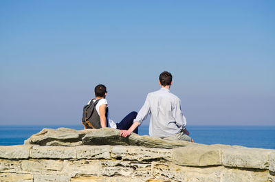 Rear view of men sitting on rock by sea against clear sky