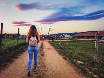 Rear view of woman walking on field against sky during sunset