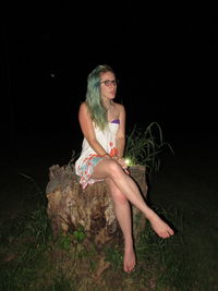 Full length portrait of young woman sitting on tree stump at night