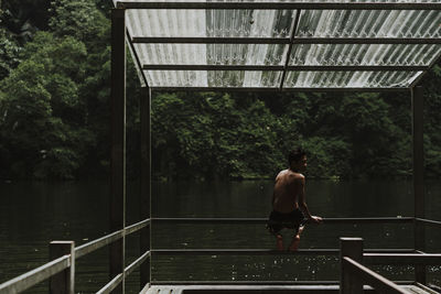 Rear view of shirtless boy sitting by lake on railing