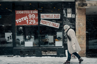 Side view of man walking on street by store during snowfall