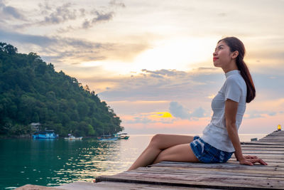 Woman sitting on shore against sky during sunset