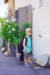 Boy with backpack stays near the wooden door in typical european small village, summer family travel