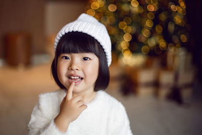 Beautiful korean child girl in in a white knitted sweater and hat stands at the christmas tree 