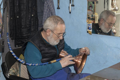 Senior man carving on brass container while sitting in workshop