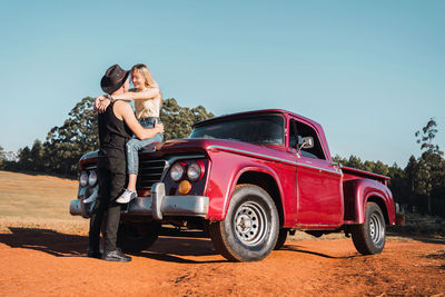 Cool boyfriend embracing smiling girlfriend sitting on hood of red retro pickup parked on sandy road