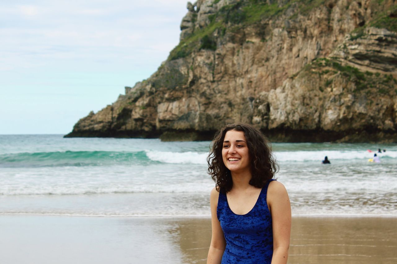 sea, beach, one person, water, real people, shore, rock - object, nature, sky, front view, smiling, looking at camera, outdoors, horizon over water, leisure activity, day, long hair, happiness, lifestyles, beauty in nature, portrait, scenics, standing, young adult, young women, people