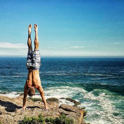 Rear view of shirtless man practicing handstand on rock at beach against sea and sky