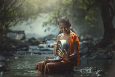 Smiling monk holding bowl and plate while sitting on rock amidst stream
