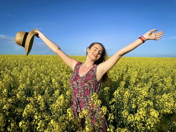 Happy young holding a straw hat in her hands while standing in a field of canola flowers