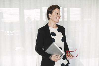 Smiling businesswoman holding eyeglasses and laptop standing at office